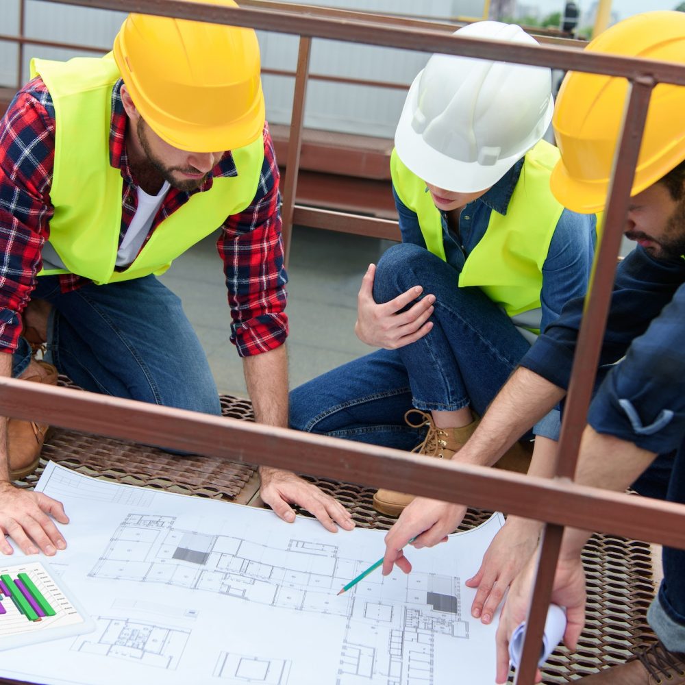 three architects in hardhats working with blueprints on roof